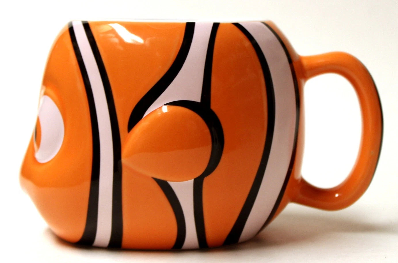 An orange ceramic mug shaped like a clownfish, featuring black and white stripes, a raised eye on the side, and a matching orange handle, shown against a plain white background.