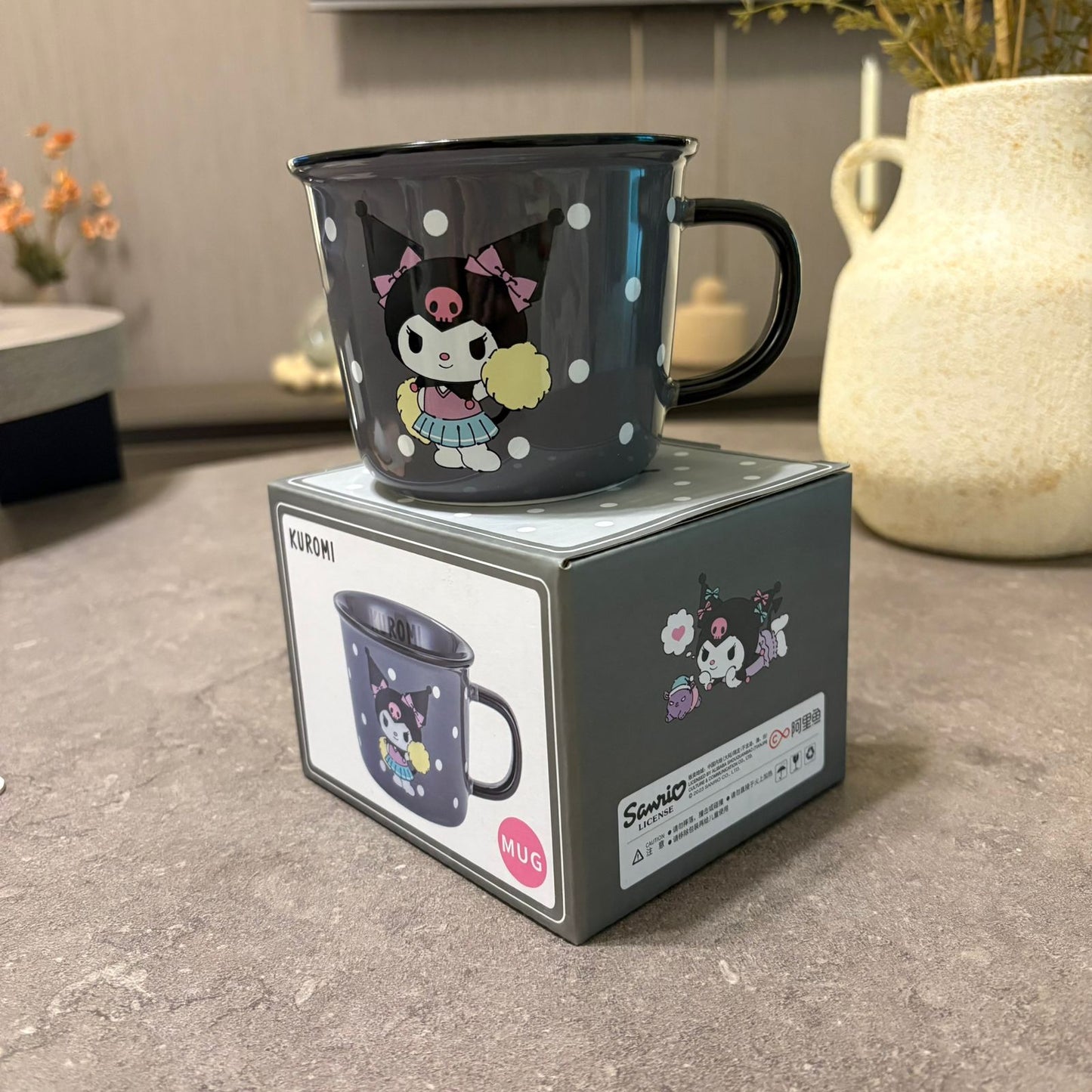 A dark gray Kuromi enamel mug with white polka dots and a black handle, sitting on top of its gray Sanrio-branded packaging box on a stone countertop.