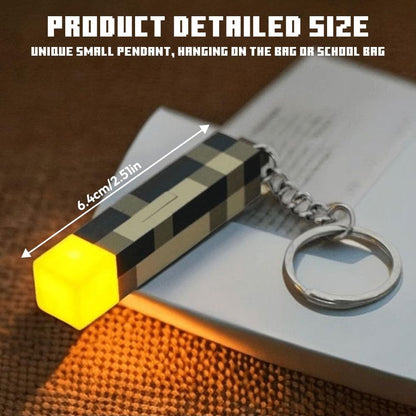 A small rectangular pendant keychain with a glowing yellow cube end, shown resting on a book. The pendant has a black, beige, and gray checkered pattern, and a silver key ring is attached.