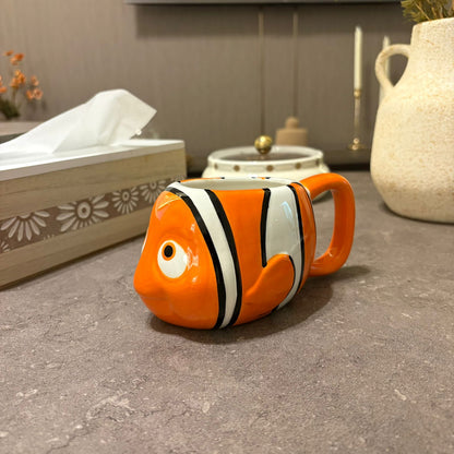 An orange and white clownfish-shaped ceramic mug with a handle, placed on a gray stone countertop in a kitchen setting.