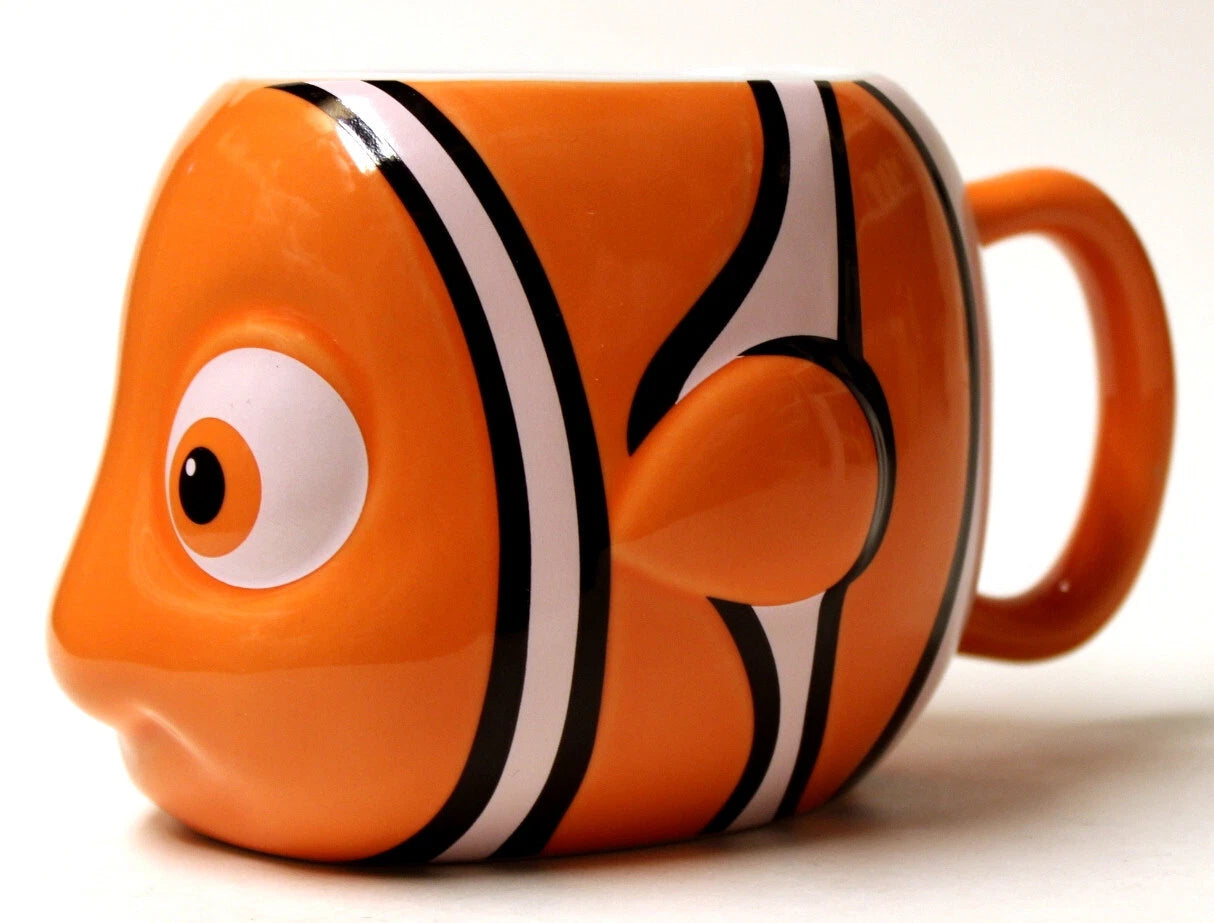 An orange ceramic mug shaped like a clownfish, featuring black and white stripes, a large cartoon eye, and a handle on the right side, photographed against a plain white background.
