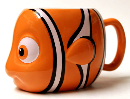 An orange ceramic mug shaped like a clownfish, featuring black and white stripes, a large cartoon eye, and a handle on the right side, photographed against a plain white background.