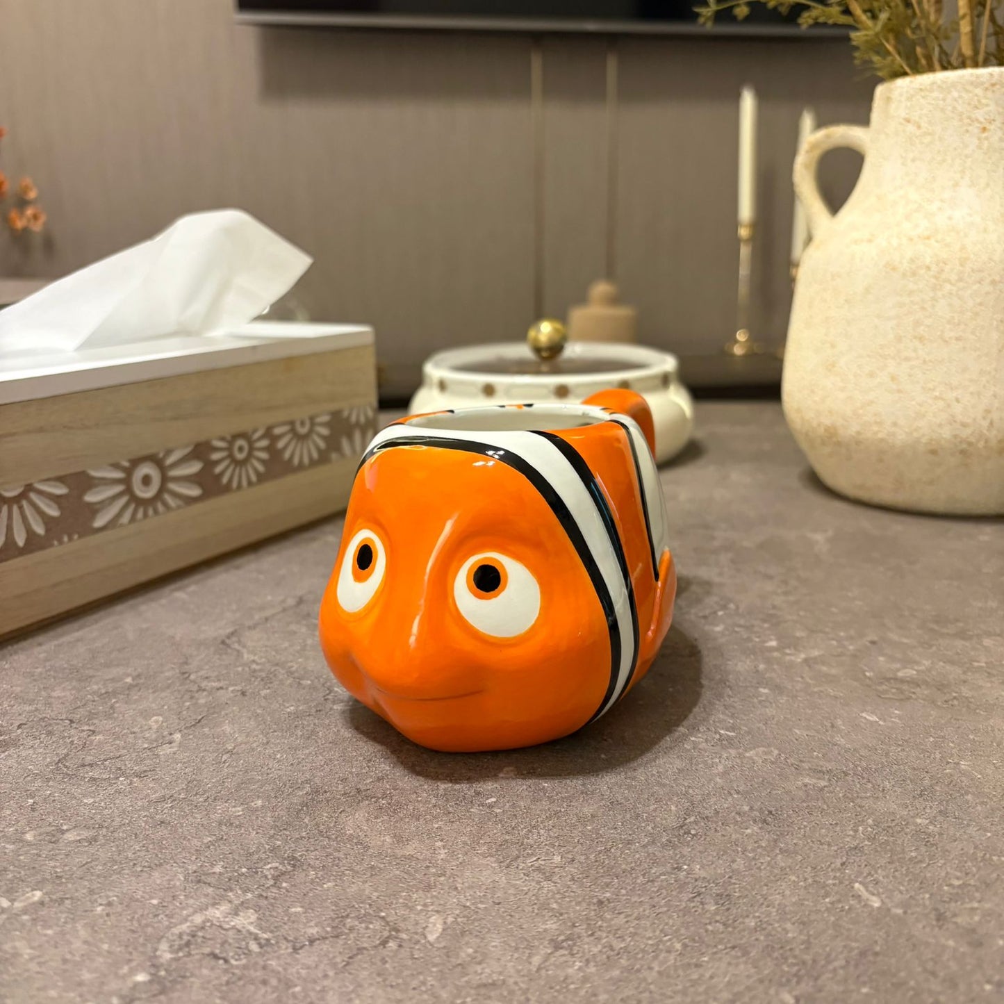 An orange clownfish-shaped ceramic tissue box cover or container placed on a gray tabletop in a home setting, with a tissue box, small white dish, and cream vase in the background.