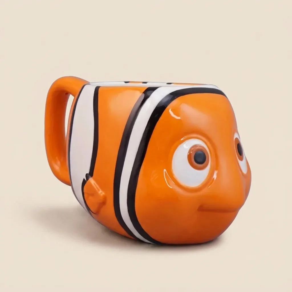 An orange clownfish-shaped ceramic mug with white and black stripes, designed to look like the character Nemo, on a solid beige background.