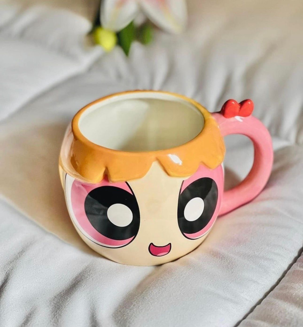 A ceramic Powerpuff-style mug with a pink handle, large cartoon eyes, and peach hair, photographed on a soft white fabric surface.