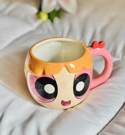 A ceramic Powerpuff-style mug with a pink handle, large cartoon eyes, and peach hair, photographed on a soft white fabric surface.
