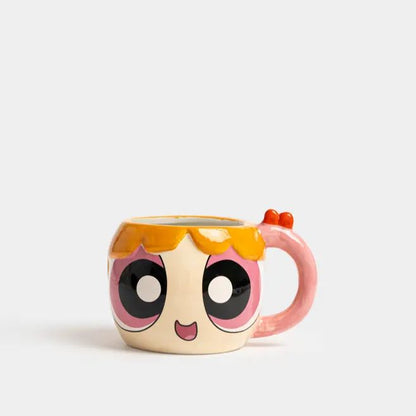 A ceramic mug shaped like a cartoon character face, featuring large black and white eyes with pink outlines, a small pink mouth, orange hair-like detailing, and a pink handle with orange accents, shown on a light gray background.