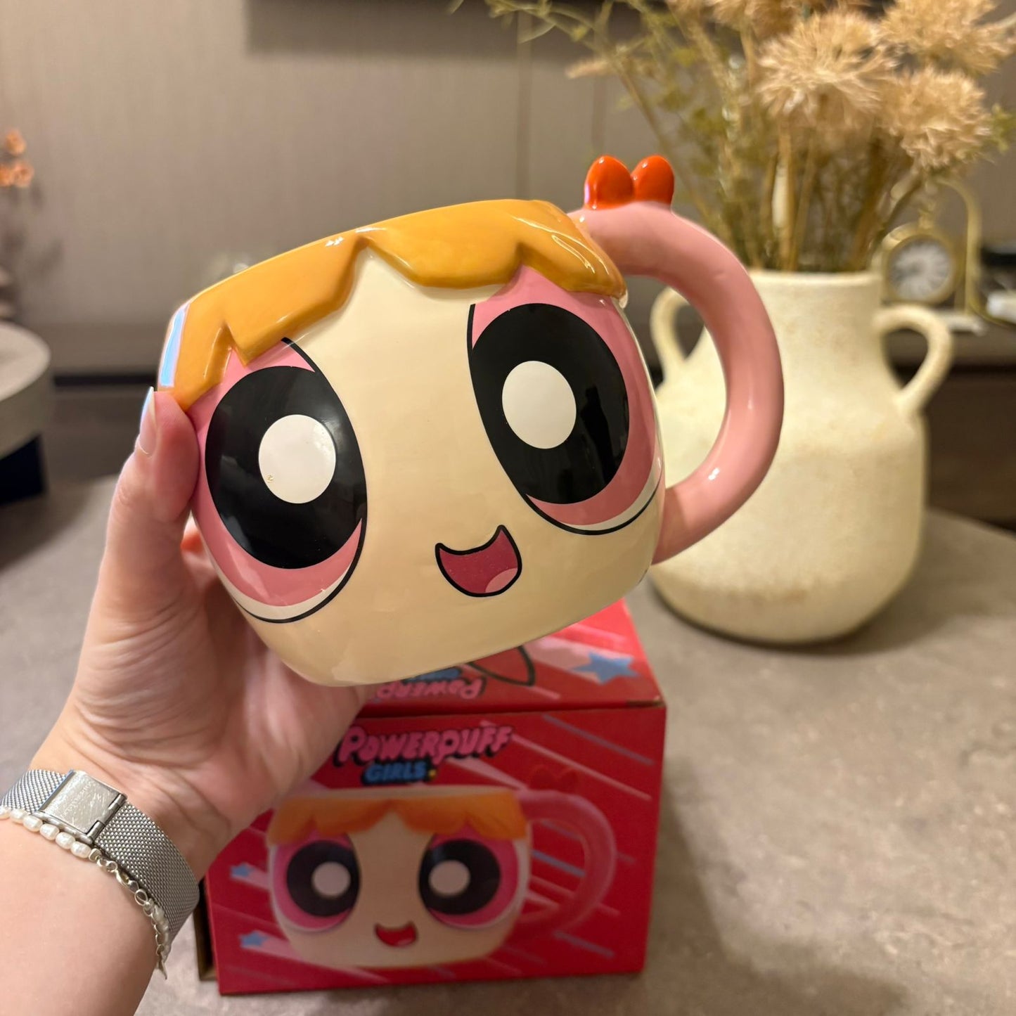 A hand holds a Powerpuff Girls ceramic mug shaped like the character Bubbles, with the red product box visible in the background on a countertop.