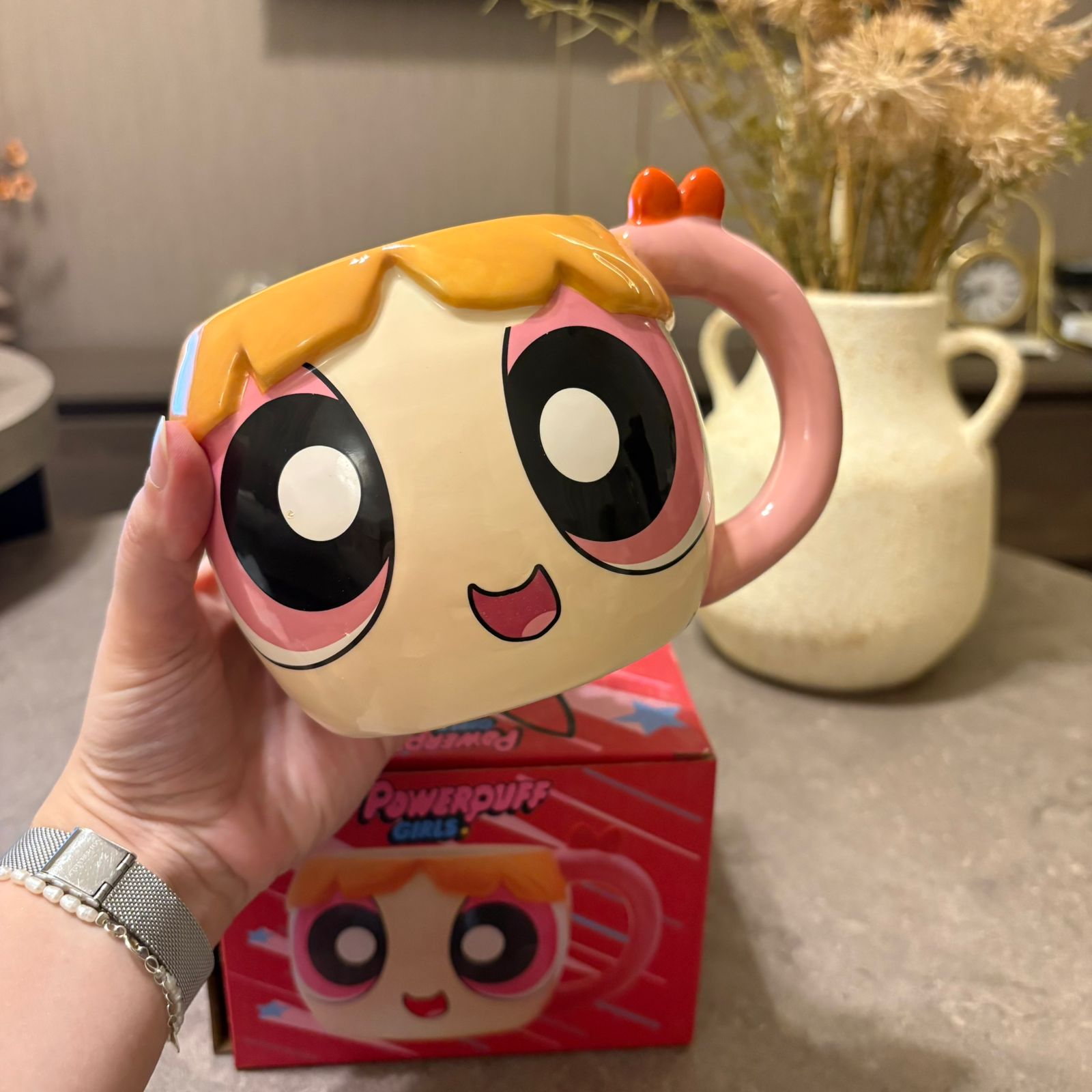A hand holds a Powerpuff Girls ceramic mug shaped like the character Bubbles, with the red product box visible in the background on a countertop.
