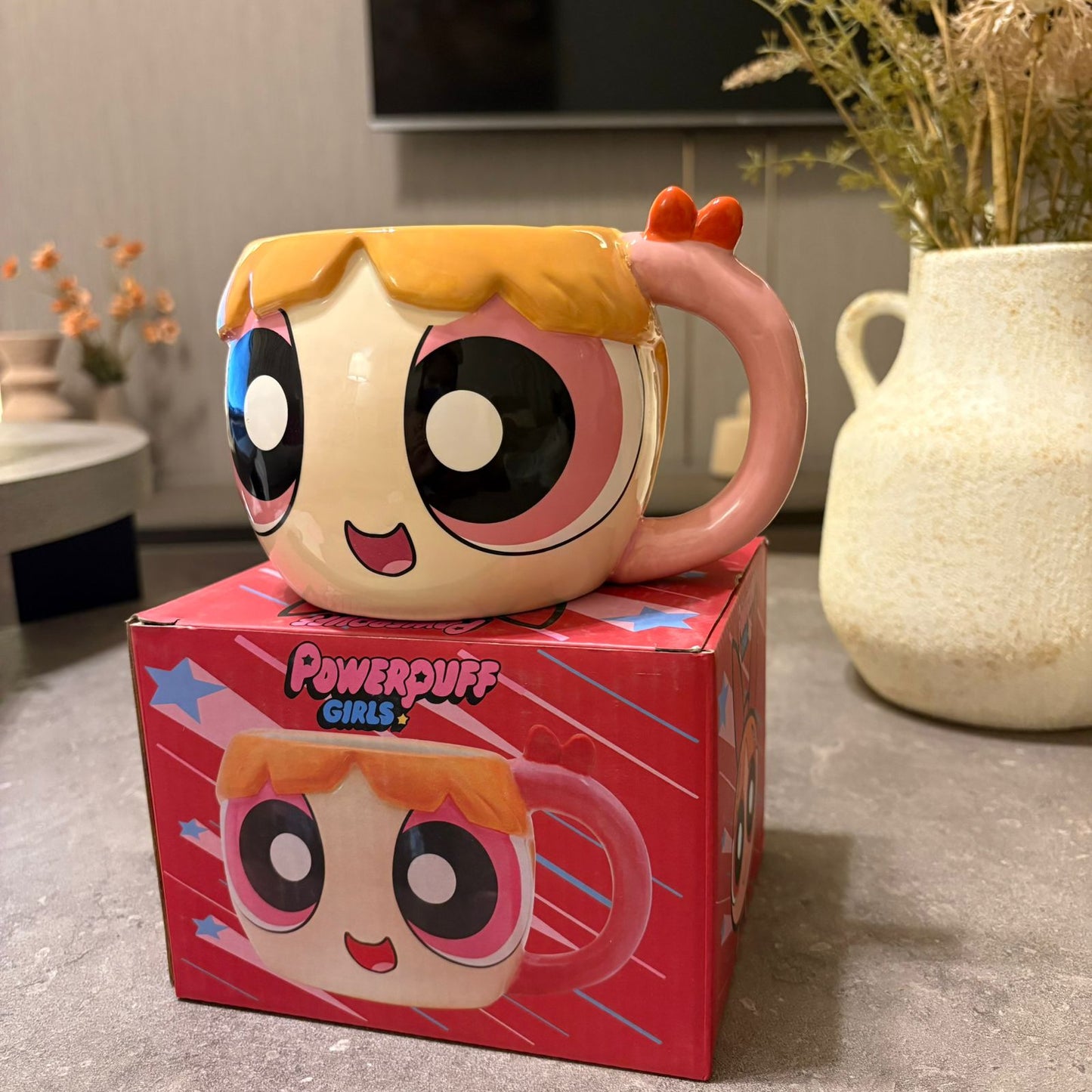 A Powerpuff Girls ceramic mug shaped like one of the characters, placed on top of its red branded box in a home setting with a vase and TV in the background.