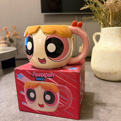 A Powerpuff Girls ceramic mug shaped like one of the characters, placed on top of its red branded box in a home setting with a vase and TV in the background.