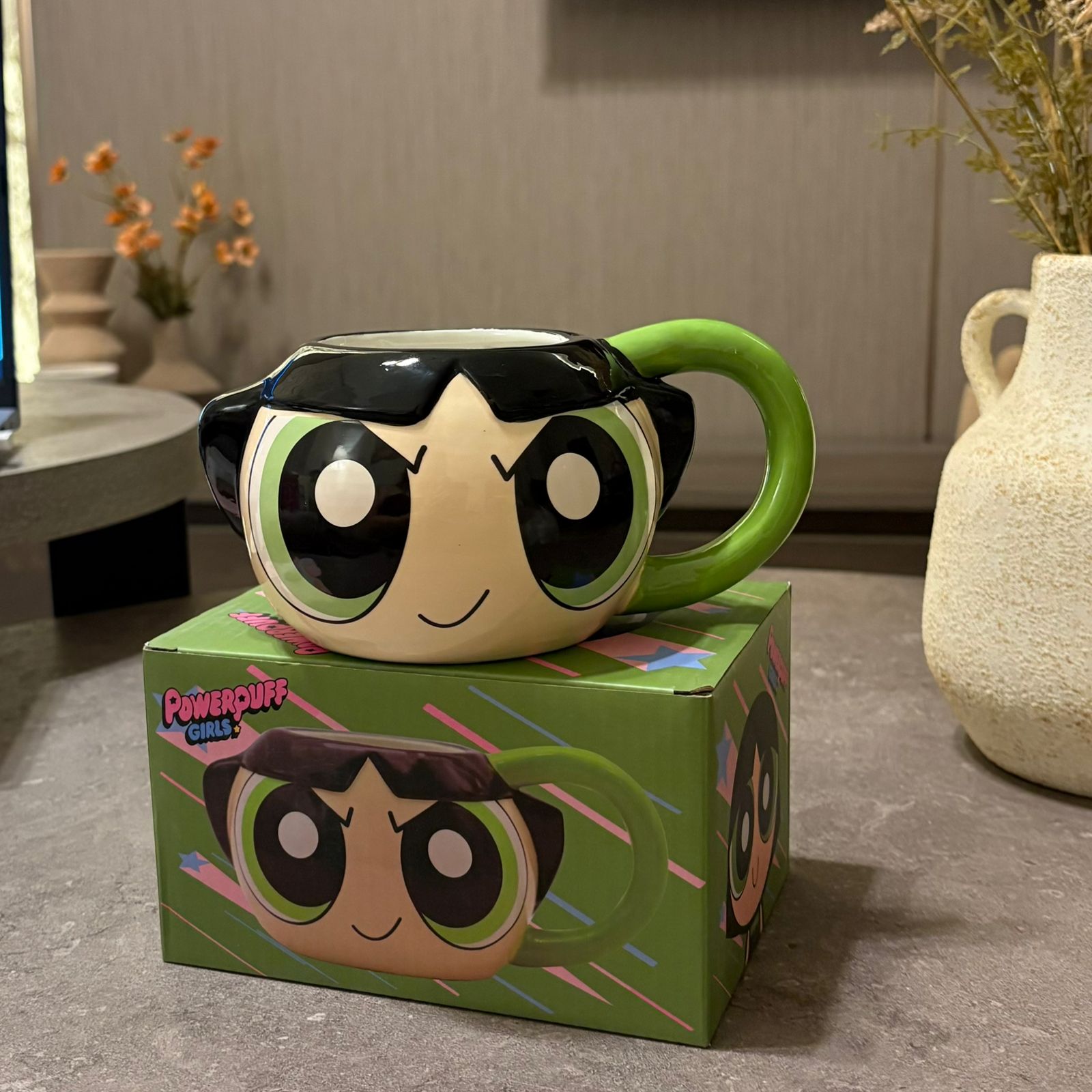 A ceramic Powerpuff Coffee mug shaped like the character Blossom, with a green handle and matching green box, set on a gray tabletop in a home environment.