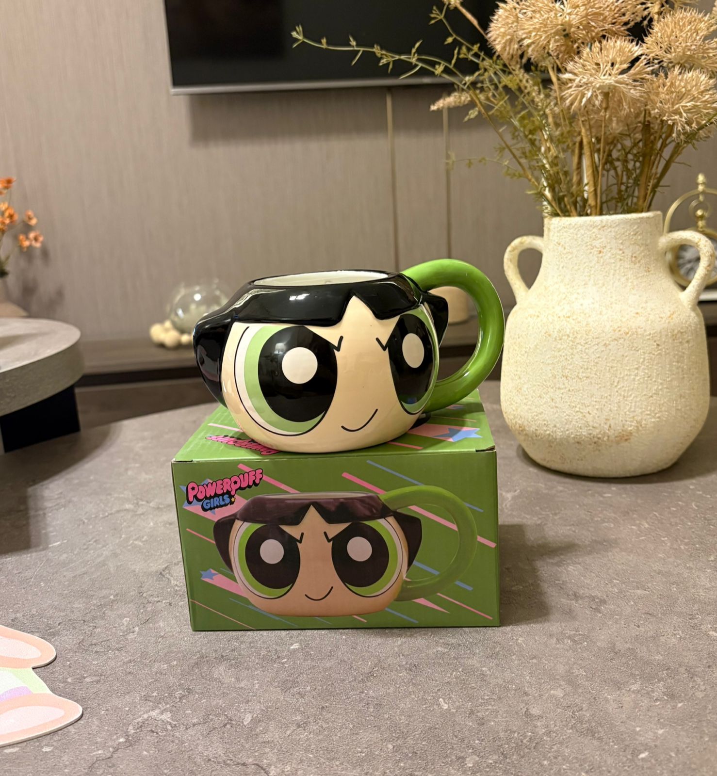 A Powerpuff Girls ceramic mug shaped like Bubbles, sitting on its green illustrated box on a gray countertop in a home setting with a vase of dried flowers in the background.