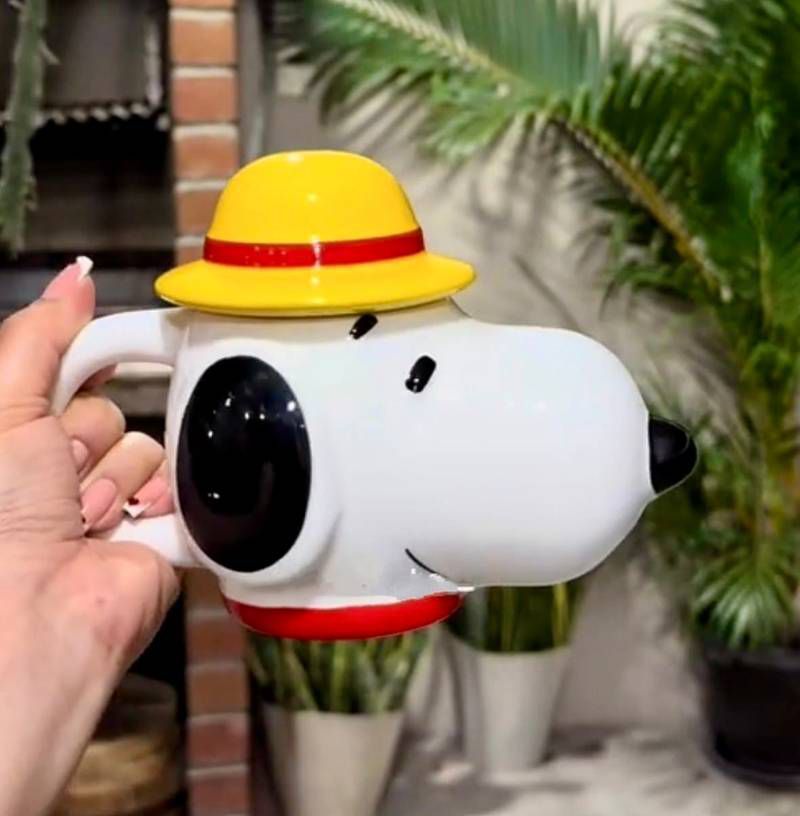 A hand holds a white ceramic mug shaped like Snoopy wearing a yellow hat with a red band, set against a background of potted plants and a brick wall.