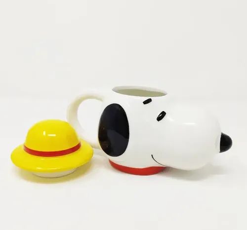 A white ceramic coffee mug shaped like a cartoon dog head with black eyes and nose, a red collar, and a small yellow hat with a red band placed beside it on a white background.