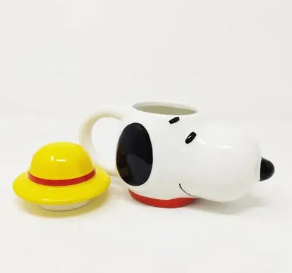 A white ceramic coffee mug shaped like a cartoon dog head with black eyes and nose, a red collar, and a small yellow hat with a red band placed beside it on a white background.