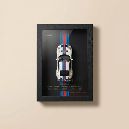 A framed wall art print featuring a top-down view of a white sports car with blue and red racing stripes on a black background, mounted on a beige wall.