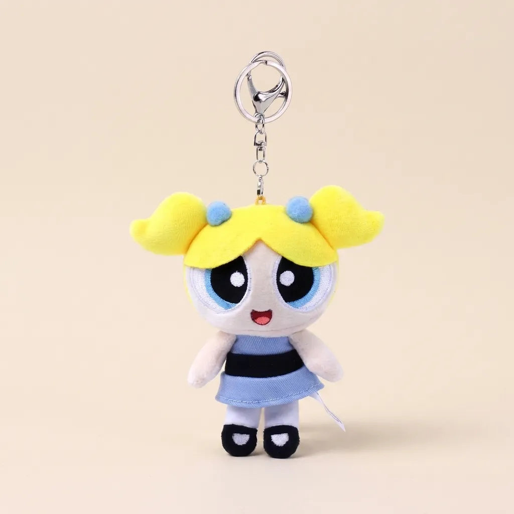 A plush keychain charm of the Powerpuff Girls character Bubbles, featuring yellow hair, a blue and black striped dress, and a silver metal key ring, set against a solid beige background.