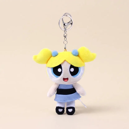 A plush keychain charm of the Powerpuff Girls character Bubbles, featuring yellow hair, a blue and black striped dress, and a silver metal key ring, set against a solid beige background.