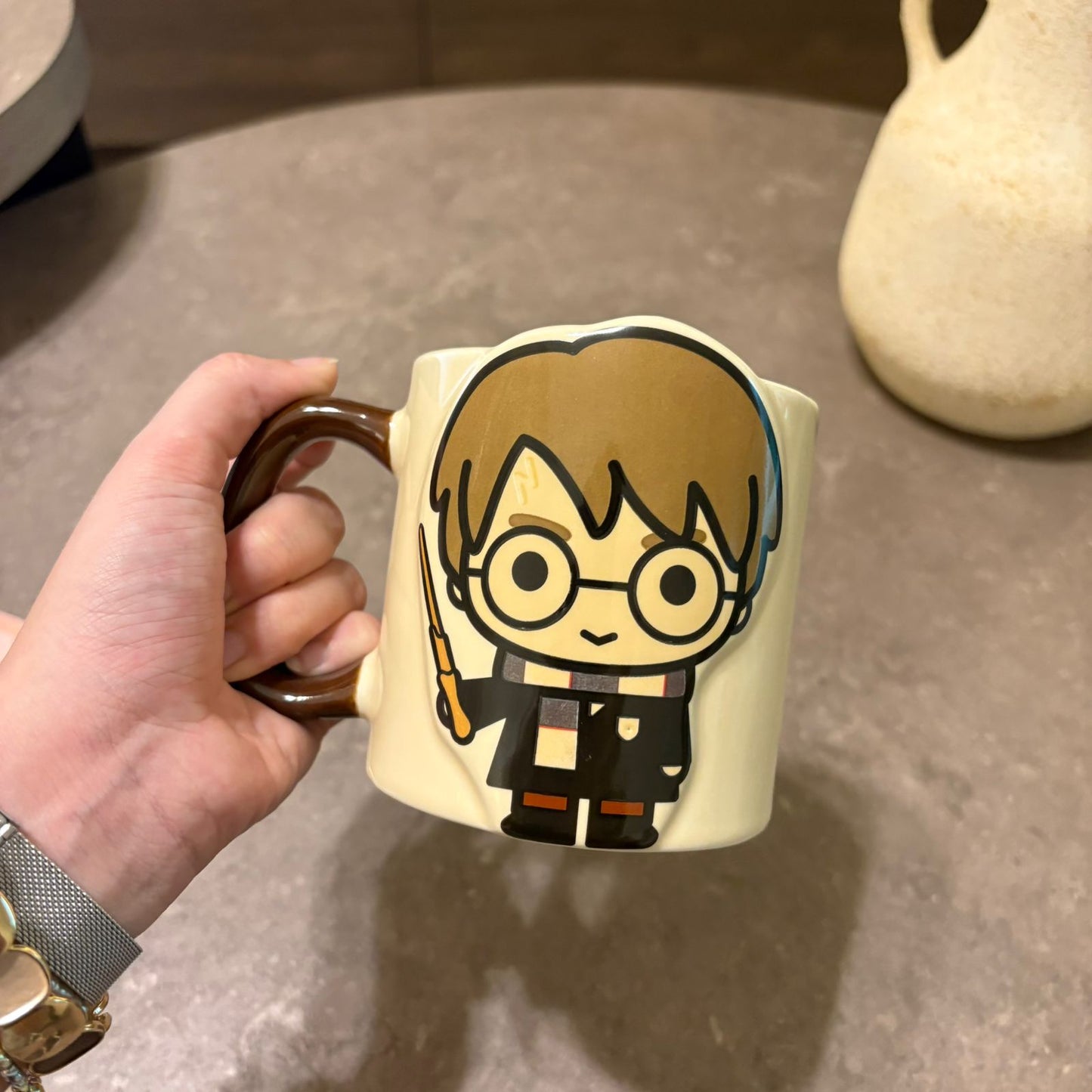 A hand holding a cream-colored ceramic mug with a cartoon character design resembling Harry Potter, set against a gray tabletop background.