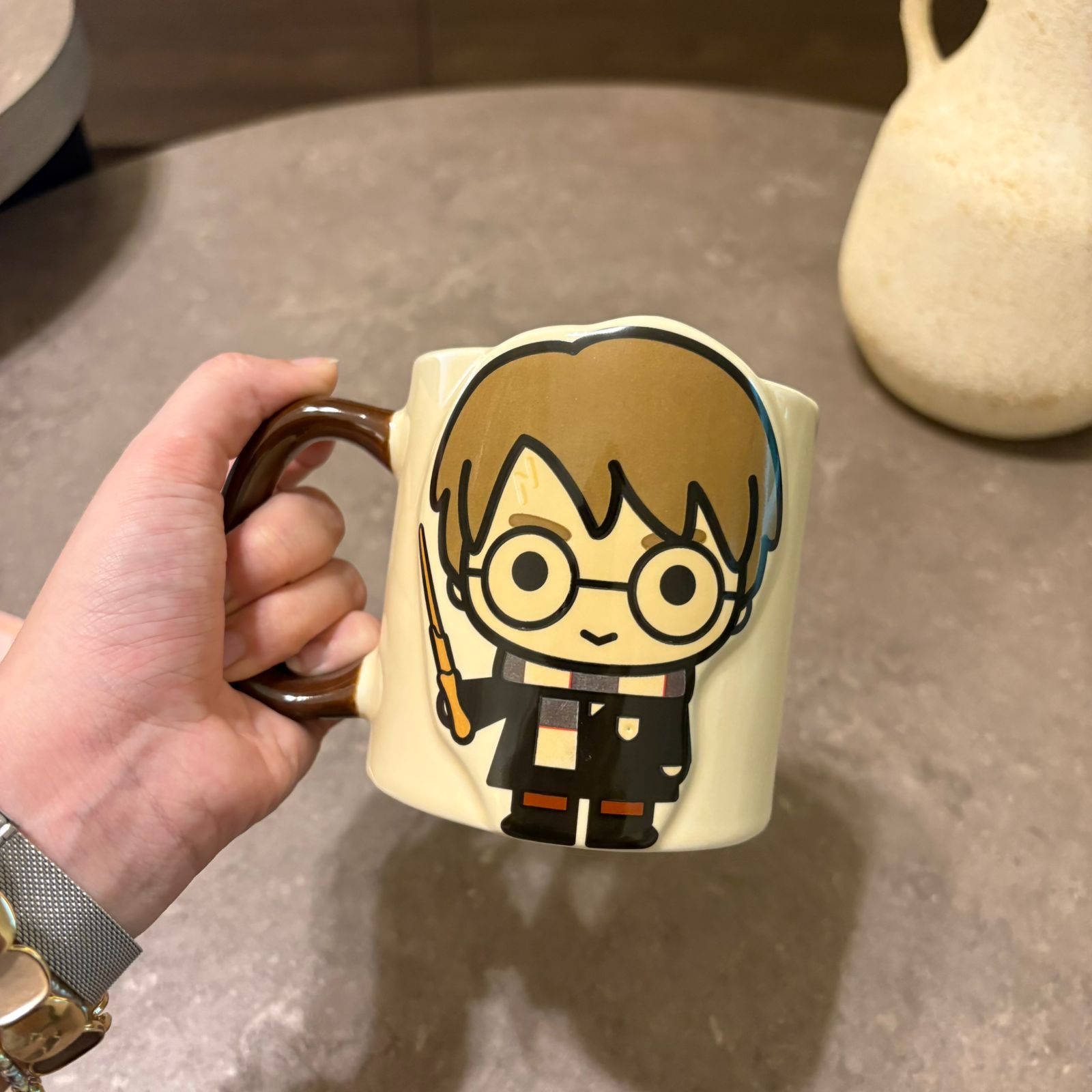 A hand holding a cream-colored ceramic mug with a cartoon character design resembling Harry Potter, set against a gray tabletop background.