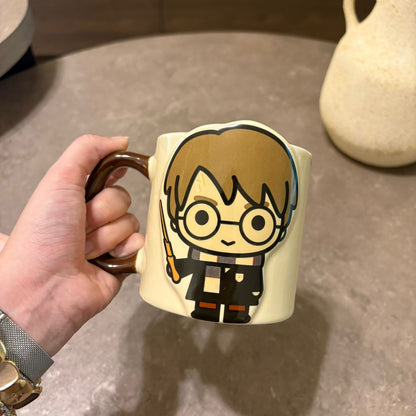 A hand holding a cream-colored ceramic mug with a cartoon character design resembling Harry Potter, set against a gray tabletop background.