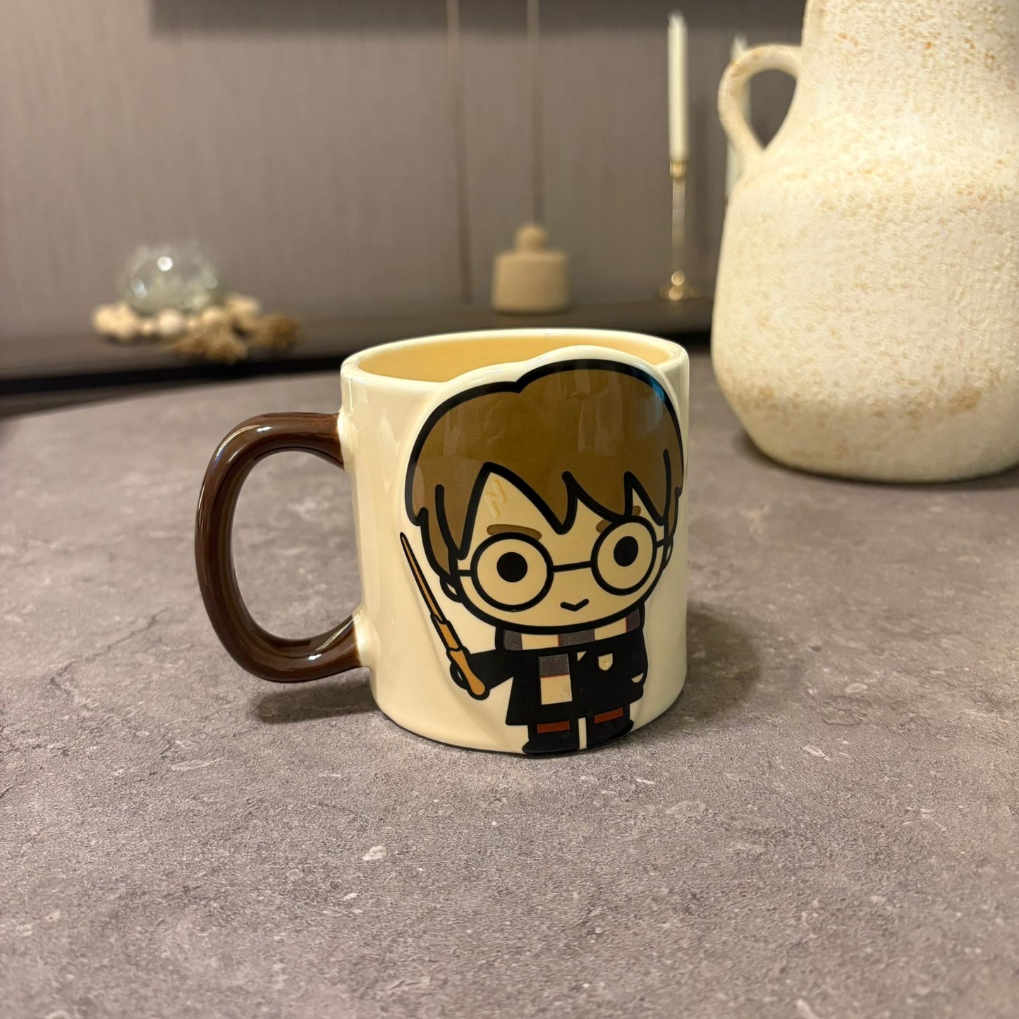 A ceramic mug featuring a cartoon illustration of Harry Potter with his wand, set on a gray stone surface with a blurred background.