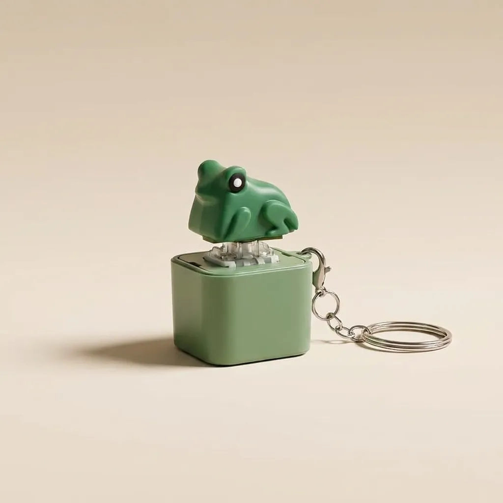 A small green frog-shaped keychain music box with a matching green cube base and silver metal keyring, photographed on a plain warm beige background.