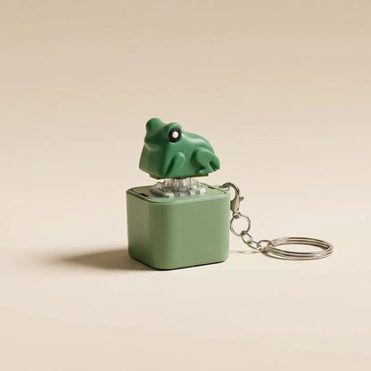 A small green frog-shaped keychain music box with a matching green cube base and silver metal keyring, photographed on a plain warm beige background.
