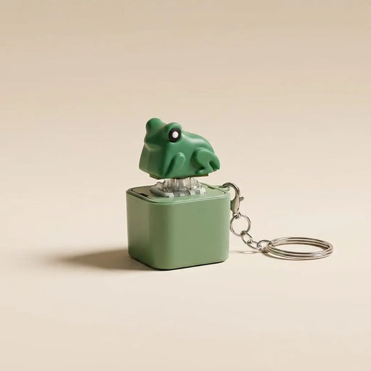 A small green frog-shaped keychain music box with a matching green cube base and silver metal keyring, photographed on a plain warm beige background.