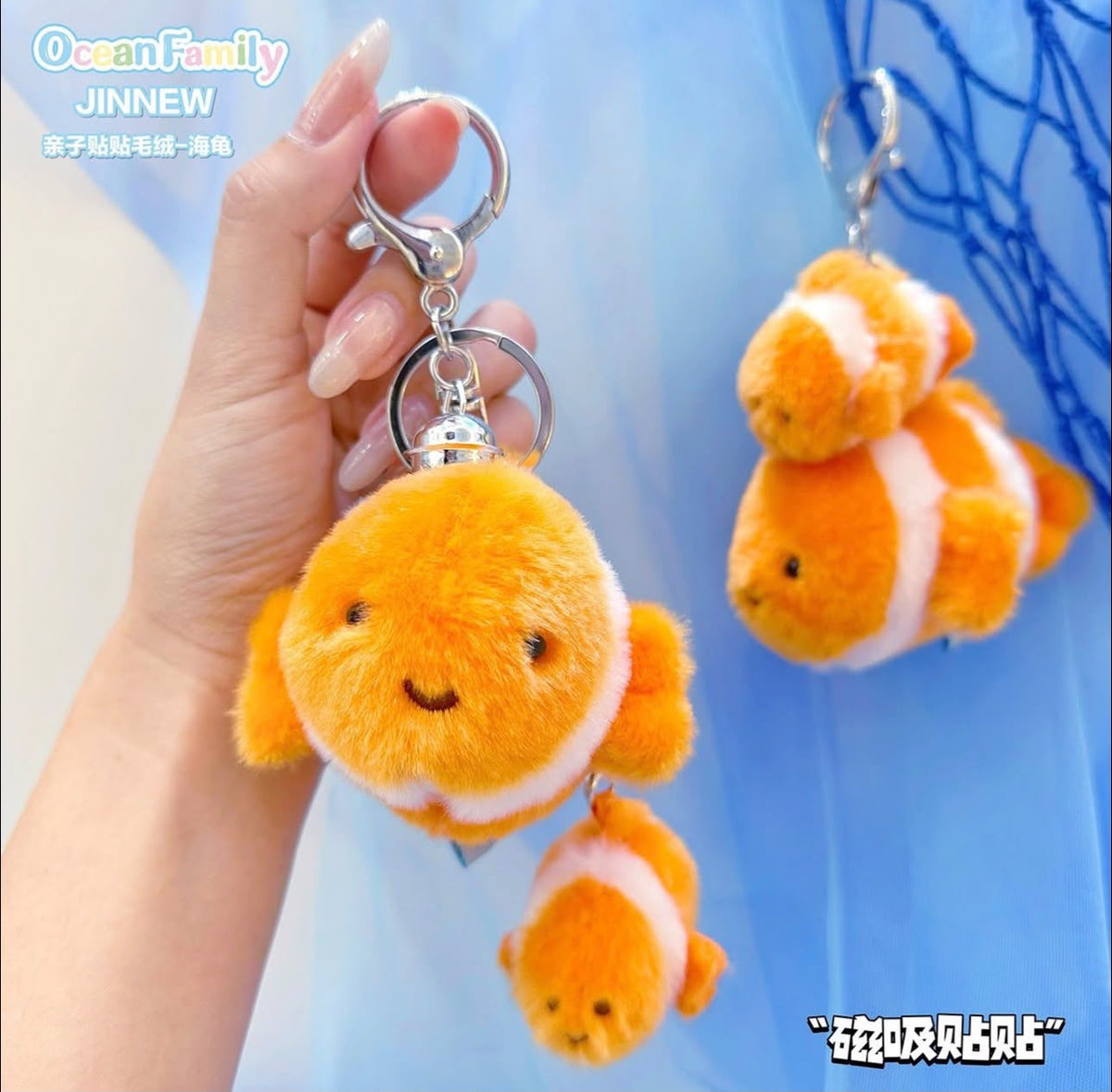 A hand holds a plush orange clownfish keychain with a silver clasp against a light blue background. A second matching plush clownfish keychain hangs on the right, and a smaller matching one is dangling below the main one.