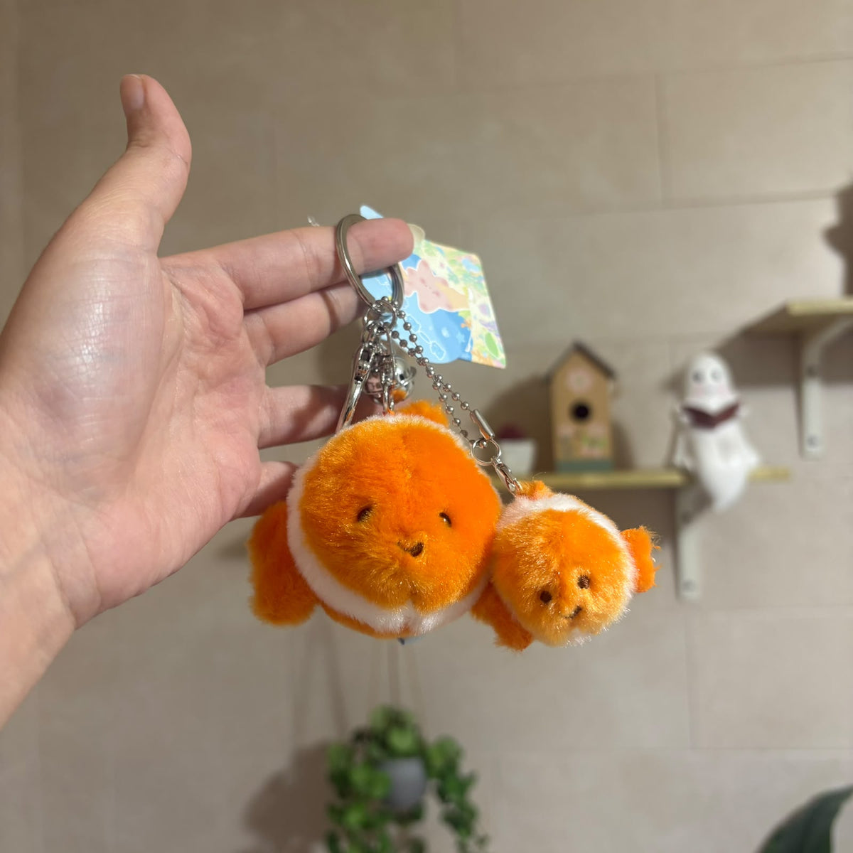 A hand holds two small orange plush fish keychains with white stripes and metal key rings, with a small map tag attached. The background shows a beige tiled wall with shelves holding small decorative items.