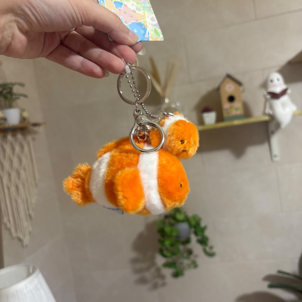 A plush fish keychain held by a hand, featuring an orange and white striped clownfish design with silver metal key rings, set against a softly blurred indoor background.