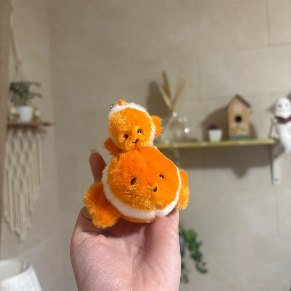 A small orange and white plush fish keychain held in a person's hand, with a softly blurred indoor background showing shelves and decor.