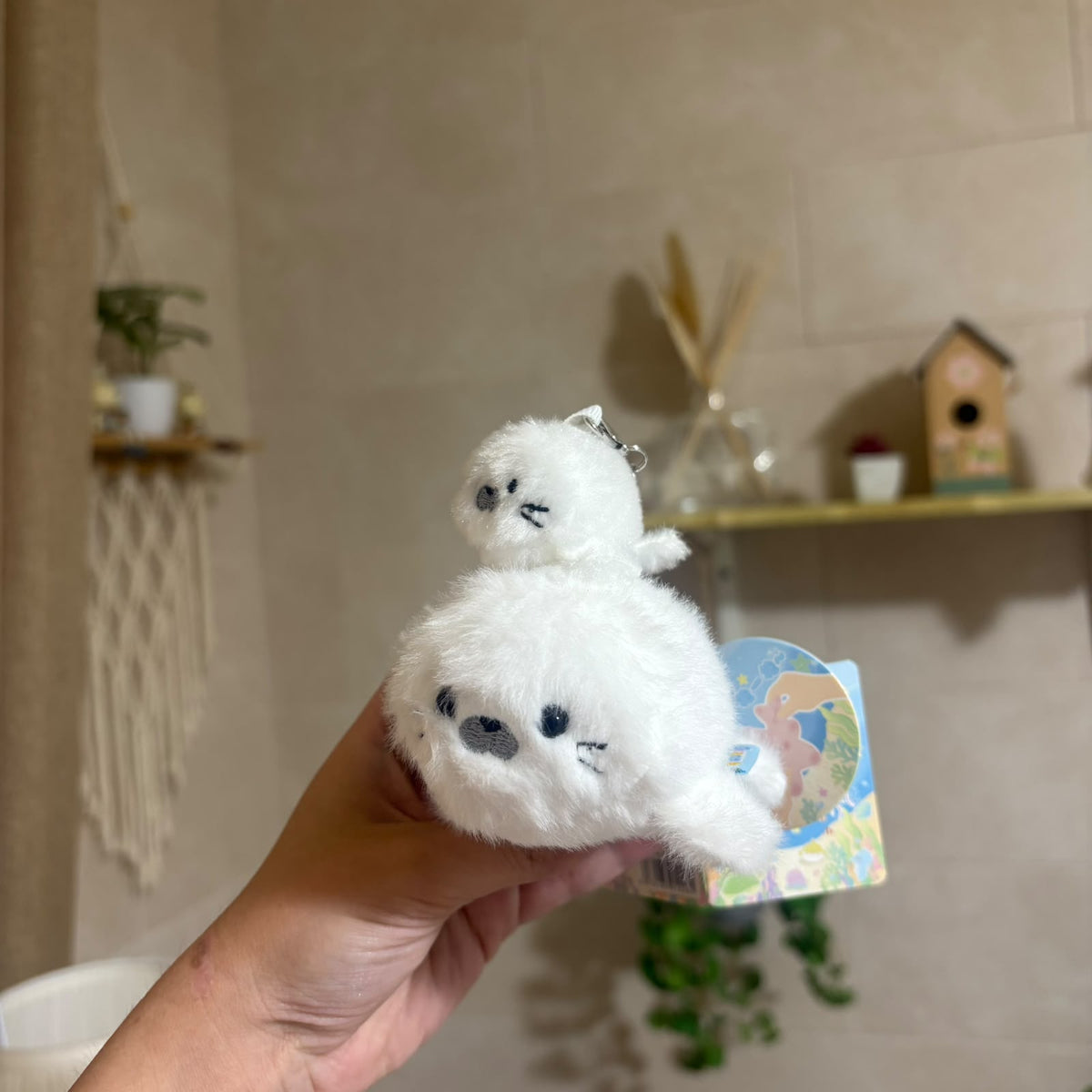 A hand holds a small white plush keychain shaped like a seal or penguin with embroidered black facial features. The plush has a small metal keyring at the top and a colorful printed backing card is attached.