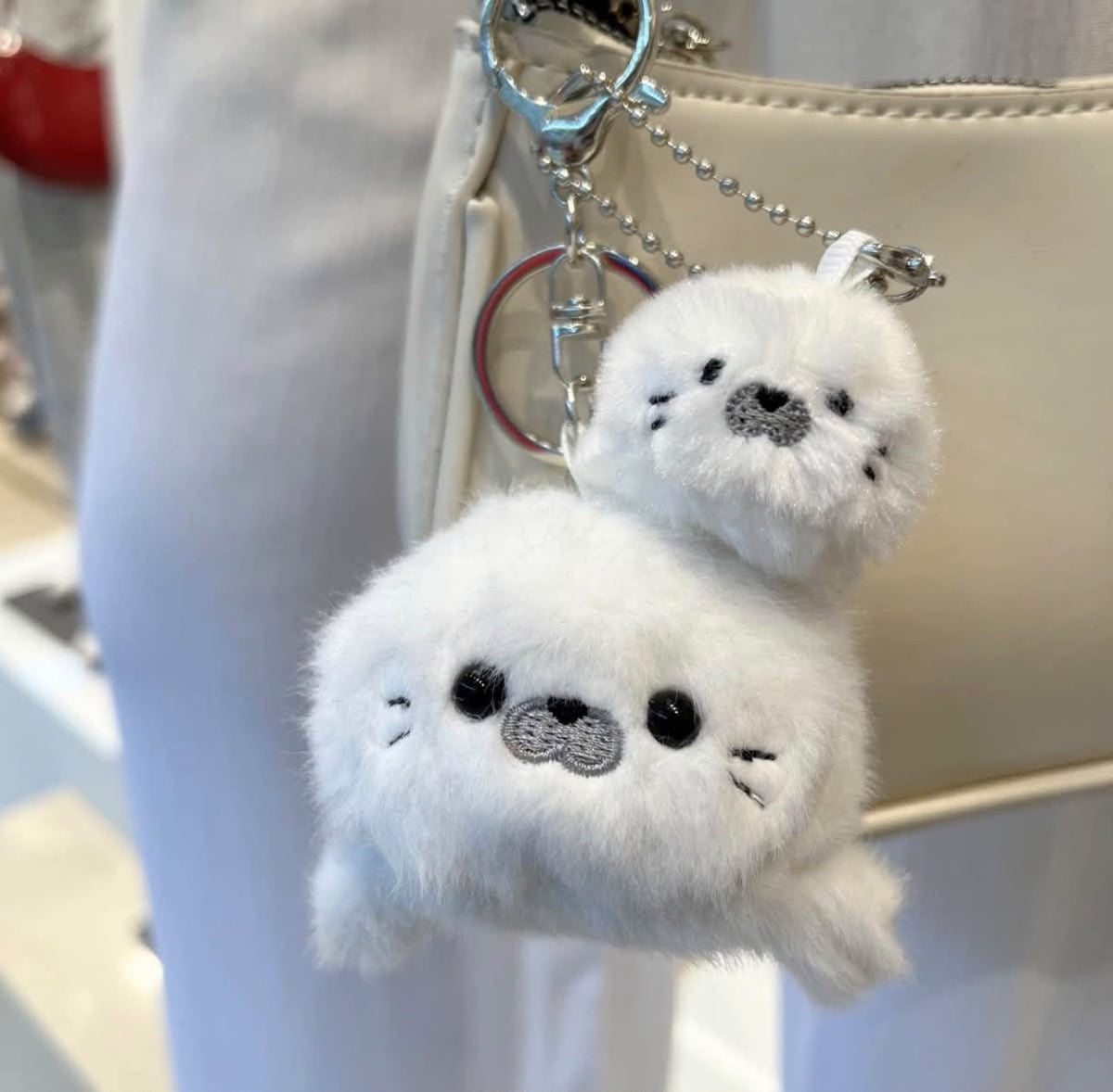 A fluffy white seal-shaped keychain charm with embroidered black and gray facial features, attached to a silver key ring and hanging near a beige handbag.