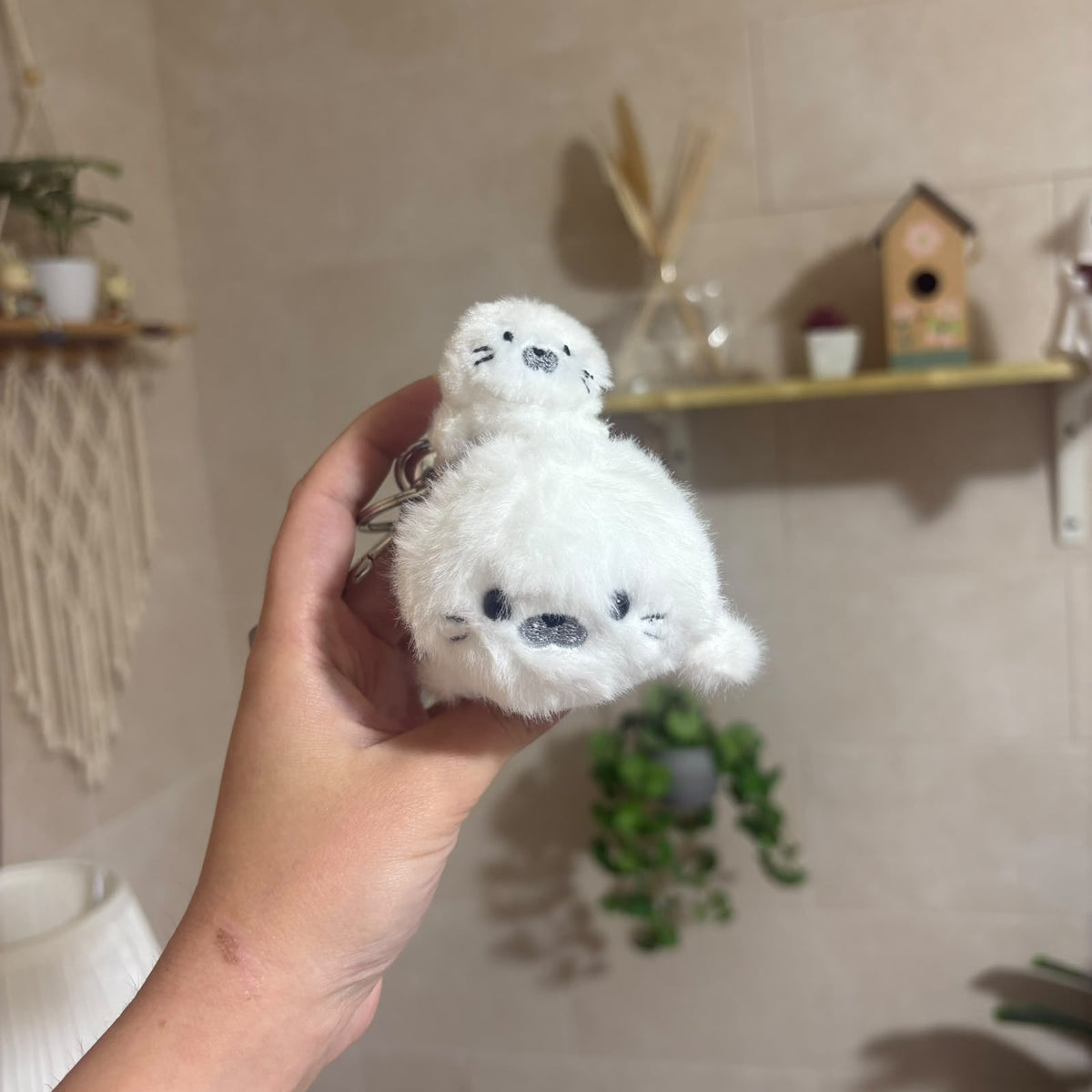 A hand holding a small white plush seal toy with embroidered black eyes and nose in a softly lit indoor setting with shelves and plants in the background.