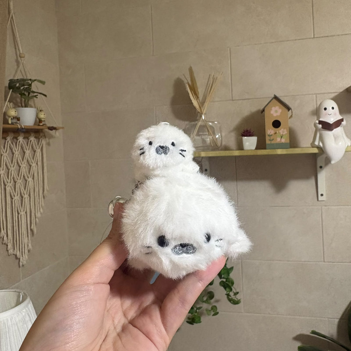 A hand holds a small white plush seal keychain in front of a beige tiled wall with shelves and decorative items in the background.