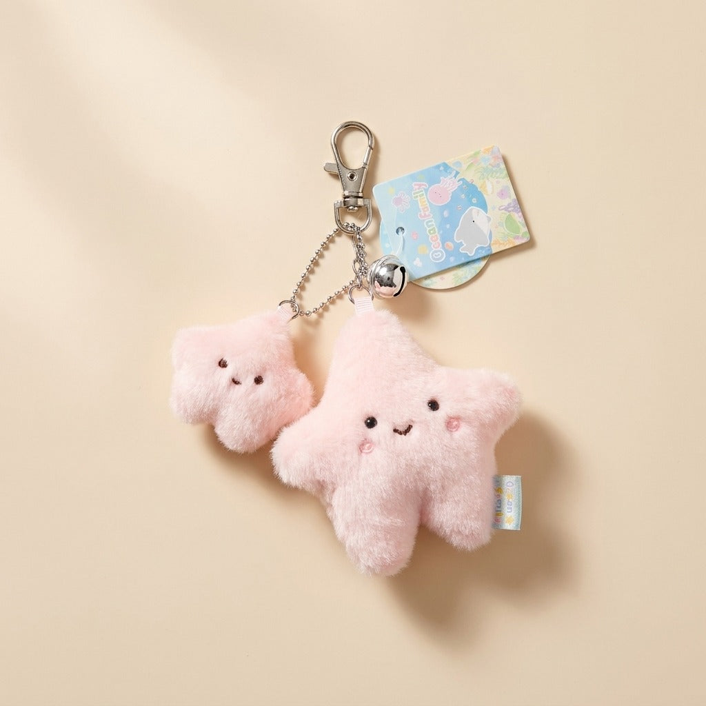 A cute pink plush star keychain set featuring two smiling star charms in different sizes, attached to a silver metal clasp and chain with a small decorative hangtag, photographed on a warm beige background.