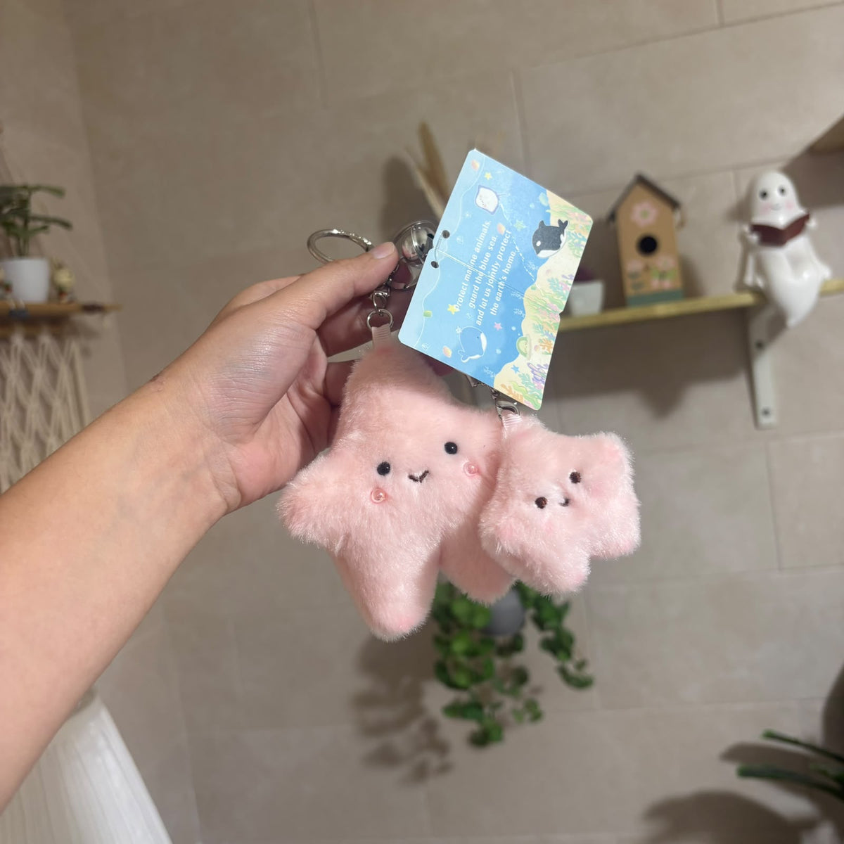 A hand holds a pink fluffy keychain featuring two cute star-shaped plush charms with embroidered faces and a blue product tag, set against a softly blurred indoor background with shelves and plants.