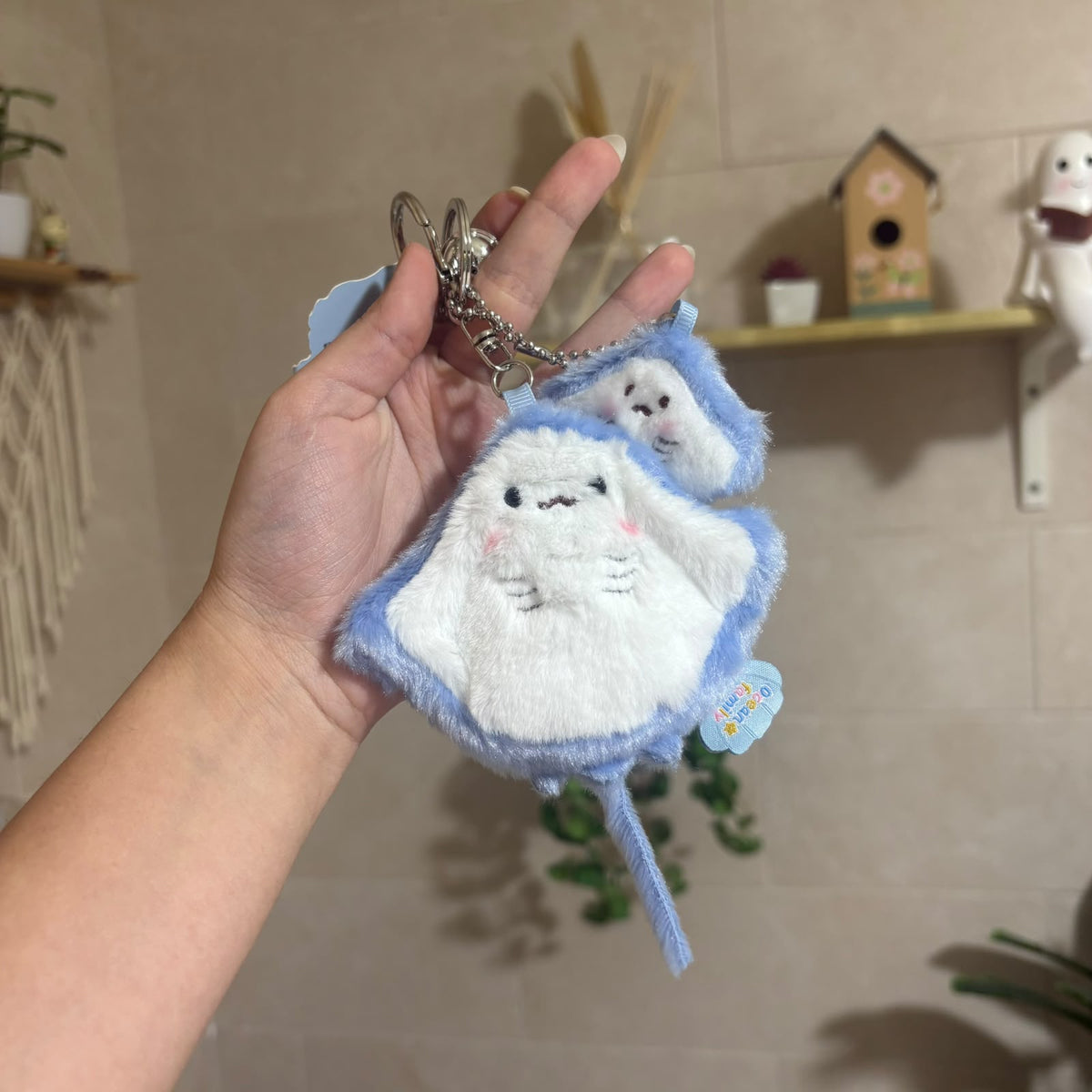 A hand holds a plush stingray-shaped keychain charm with a silver split ring. The charm is white with light blue fuzzy edges, embroidered facial features, and a small blue hanging tag.