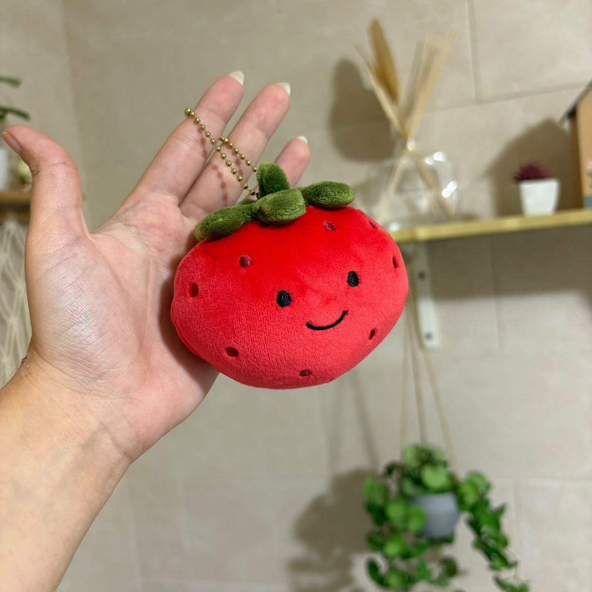 A red plush strawberry keychain with a smiling face and green stem, held in a person's hand. The plush has embroidered black eyes and mouth details and is attached to a gold ball chain.