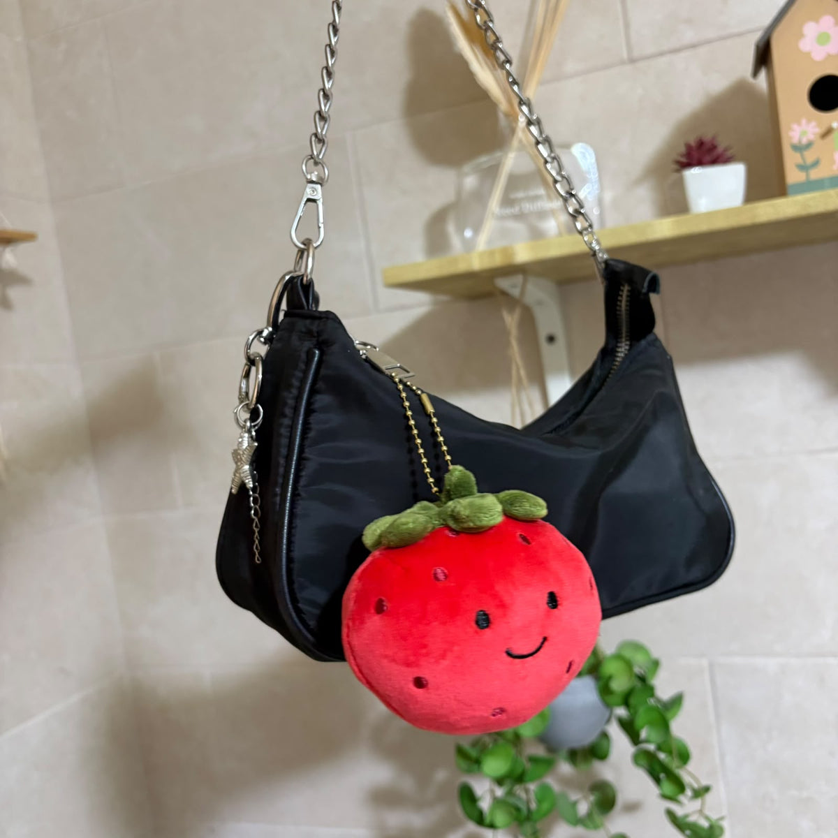 A black nylon shoulder bag with a silver chain strap, featuring a cute red strawberry plush keychain charm with green leaves and a smiling face.