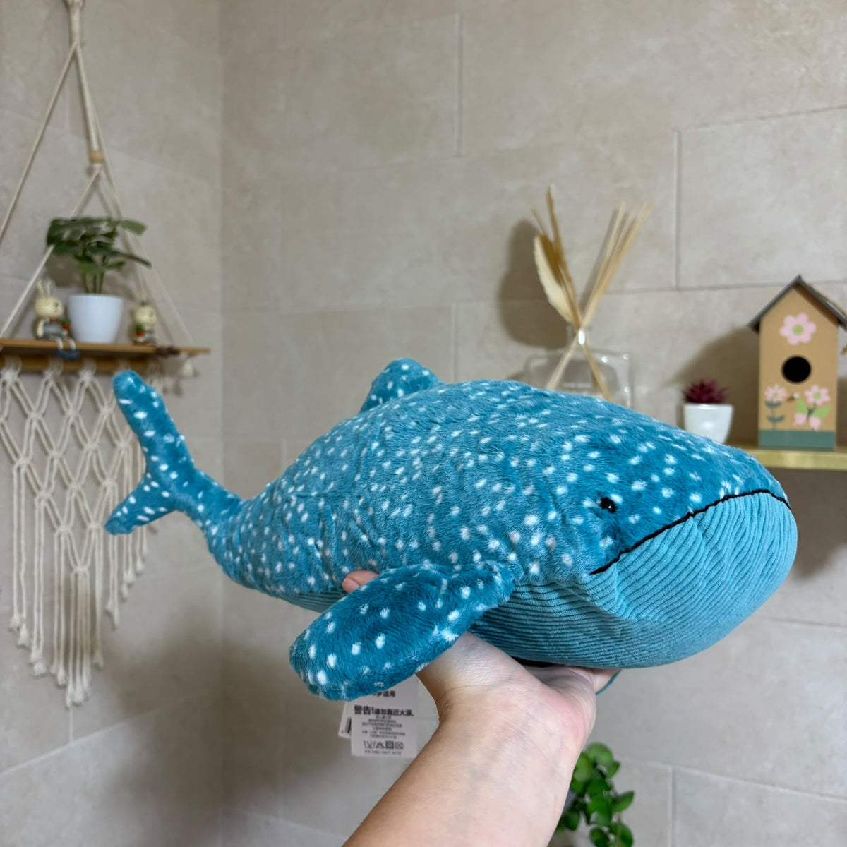 A plush whale shark toy being held up by a person's hand in a home setting with beige tiled walls, a macrame wall hanging, and small decor items in the background.