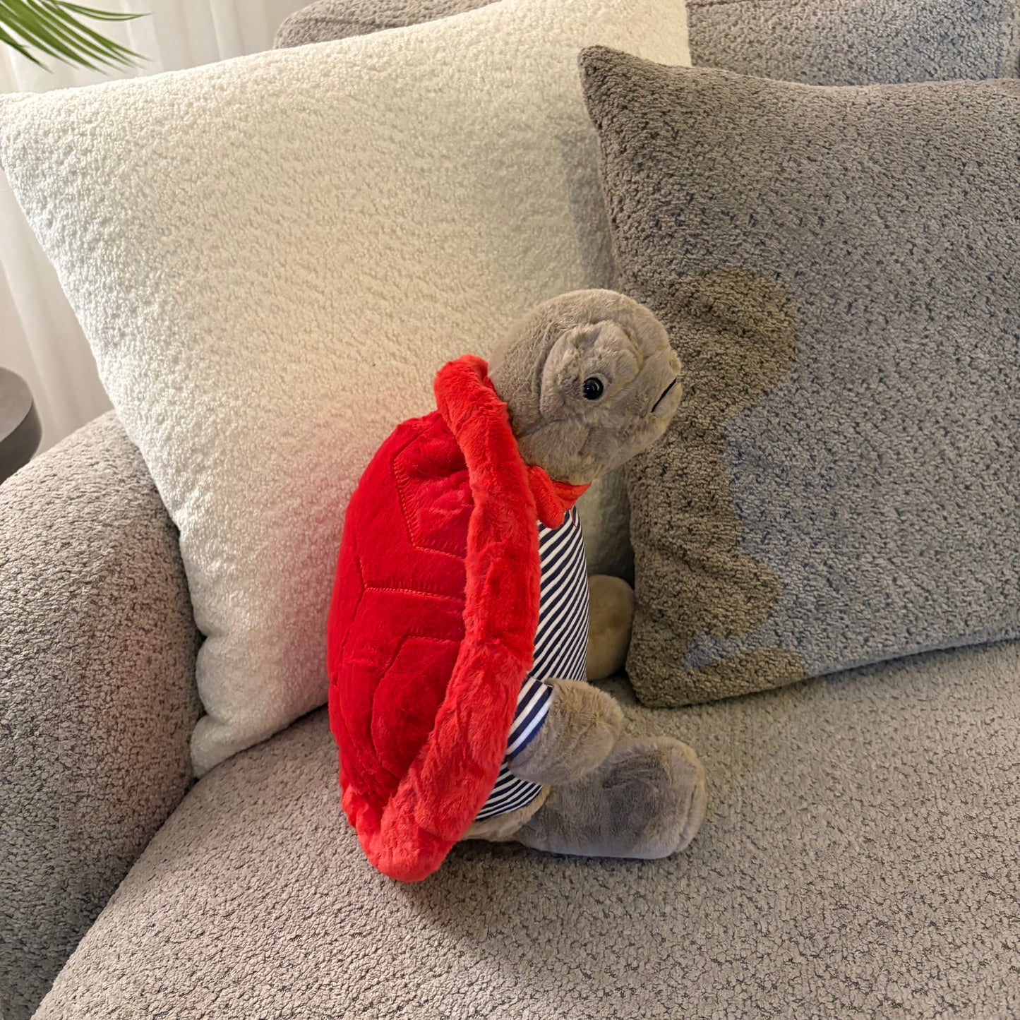 A plush stuffed turtle toy sitting on a textured gray sofa between two pillows. The turtle has a bright red shell and wears a small striped shirt.