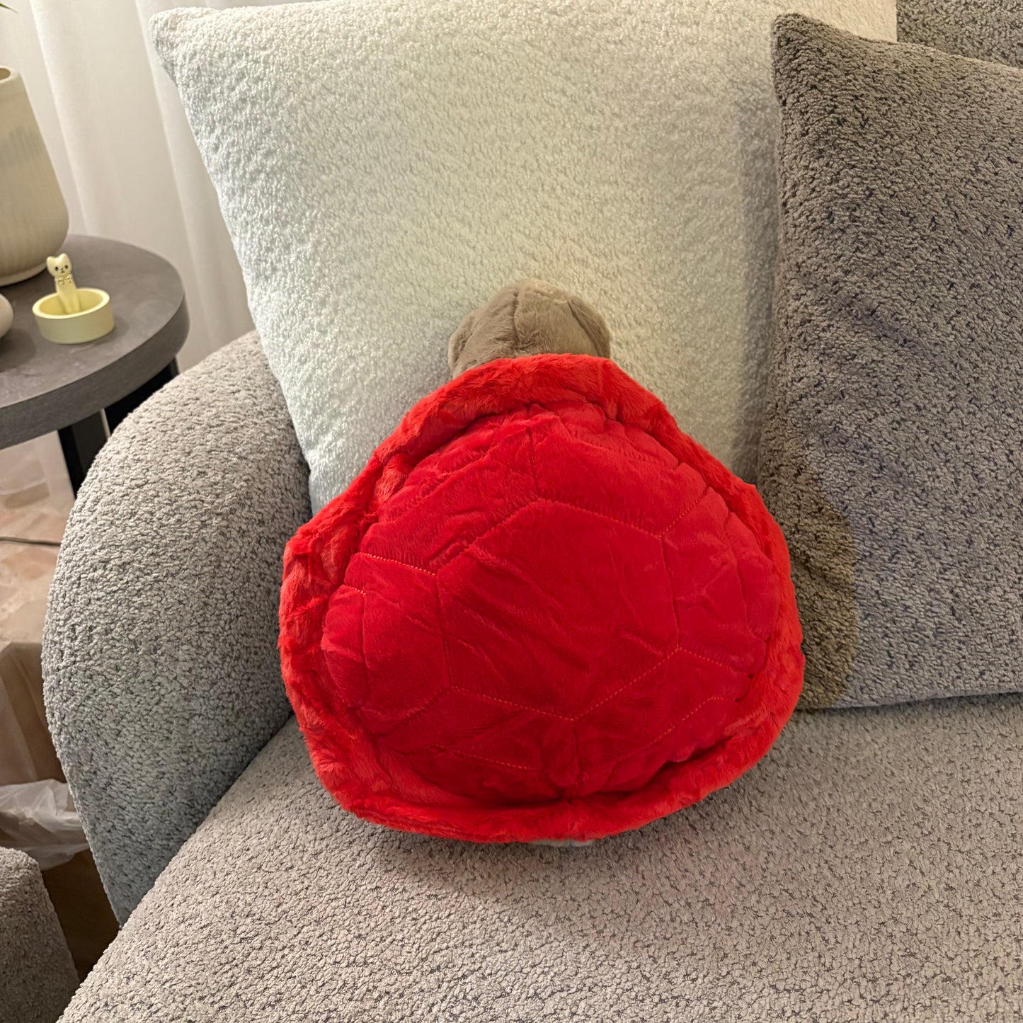 A red turtle-shaped plush pillow with a gray head resting on a gray textured sofa next to white and gray pillows.