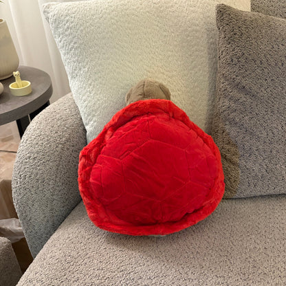 A red turtle-shaped plush pillow with a gray head resting on a gray textured sofa next to white and gray pillows.