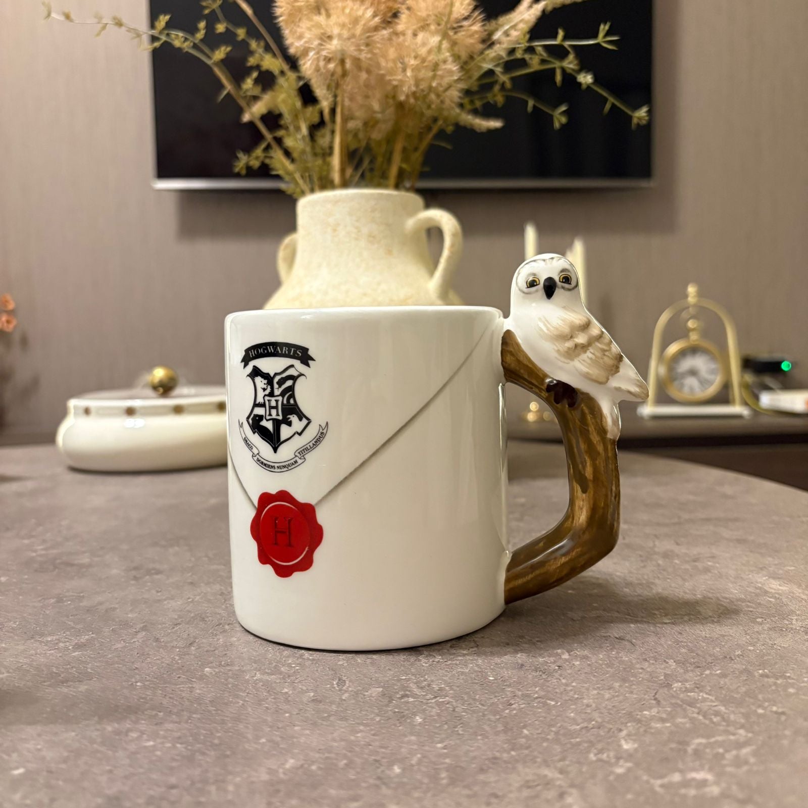 A white ceramic mug designed to look like a Hogwarts letter envelope with a red wax seal, featuring a brown owl handle perched on a branch. The mug is set on a gray tabletop with a blurred background containing a vase of dried grass and a clock.