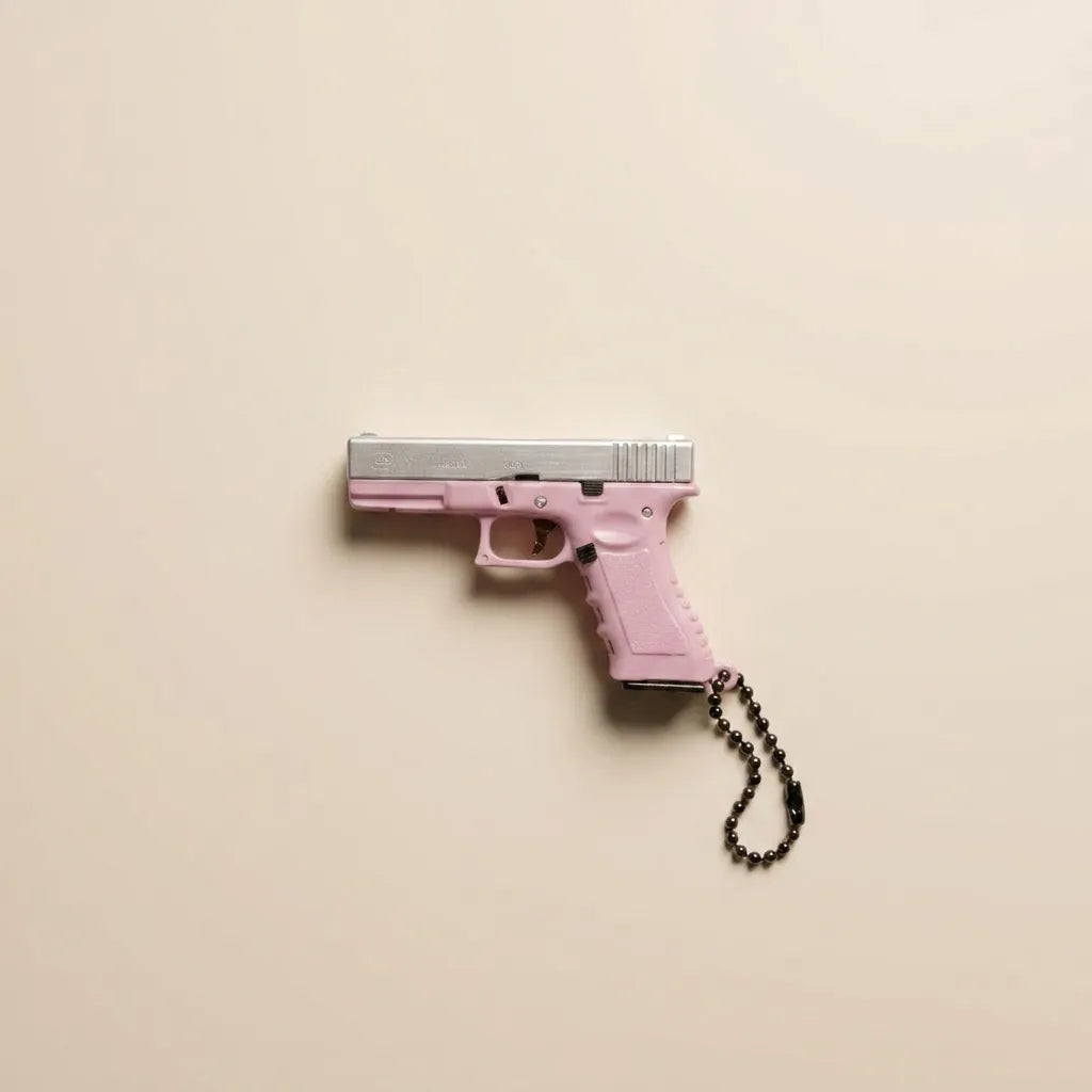 A pink handgun-shaped keychain or accessory with a silver slide and a black beaded chain, photographed against a plain warm beige background.