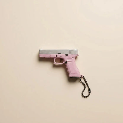 A pink handgun-shaped keychain or accessory with a silver slide and a black beaded chain, photographed against a plain warm beige background.